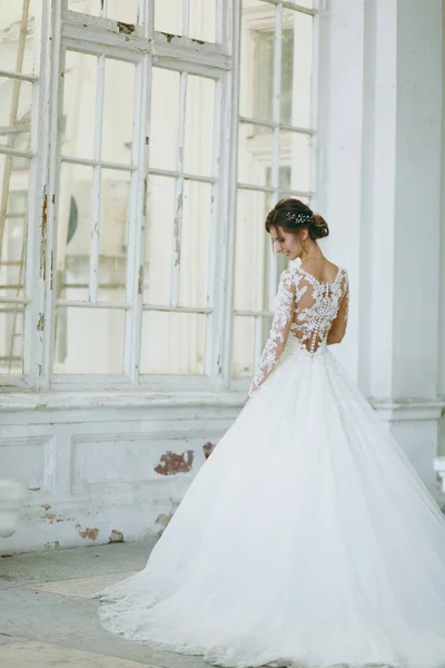 Beautiful wedding photosession. Young bride in a white lace dress with a long plume with an exquisite hairstyle in vintage interior on the veranda of an old house with columns near the garden — Stock Photo, Image