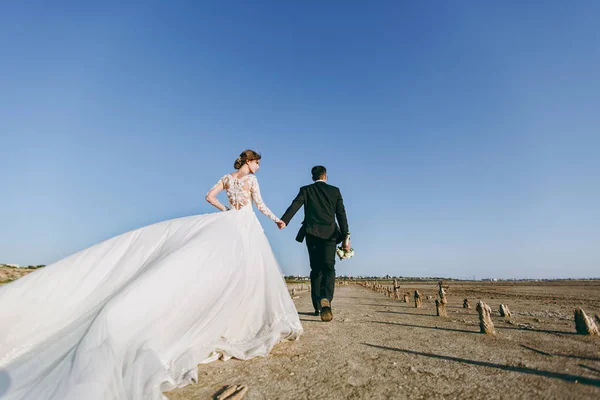 Bela sessão de casamento. Bonito noivo não raspado em uma calça preta com buquê e jovem noiva bonito em vestido padrão de renda branca com penteado requintado no passeio ao longo da costa — Fotografia de Stock