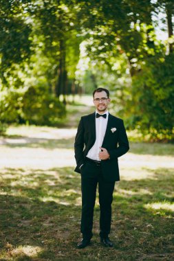 Güzel düğün photosession. Siyah bir takım elbise, beyaz bir gömlek, bir papyon ve weathery güneşli günde büyük bir yeşil bahçe çiçek dolu bir yaka çiçeği ile gözlük genç gülümseyen tıraşsız damat