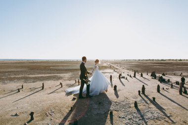 Güzel düğün photosession. Beyaz dantel desen elbise yürüyüşe kıyı şeridi boyunca mükemmel saç modeli ile sevimli gelin buketi ve genç bir siyah pantolon yakışıklı tıraşsız damat