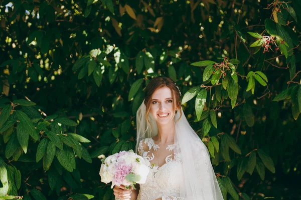 Schöne Hochzeitsfotosession. junge zarte lächelnde Braut in elegantem Spitzenkleid mit langen Ärmeln, Schleier und Strauß weißer und pinkfarbener Blumen inmitten der Zweige des grünen Busches im Garten — Stockfoto