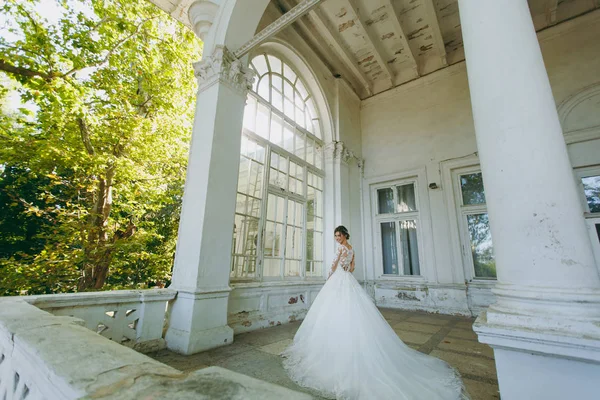 美丽的婚礼 photosession。年轻的新娘在白色花边礼服与长羽与精致的发型在老式室内的阳台上的老房子与花园附近的柱子 — 图库照片