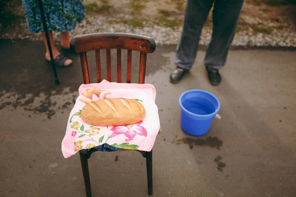 Tradiciones de la boda Compra de novia Pan pan en una toalla brillante en una silla de madera marrón contra el fondo de los pies de los ancianos y un cubo de agua al aire libre — Foto de Stock