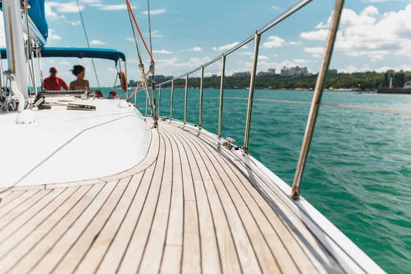 Sailing boat deck on a white yacht with teak wooden deck, metal railing, set of red ropes with people on board in the sea near the coastline in the background blue sky with clouds in sunny day — Stock Photo, Image