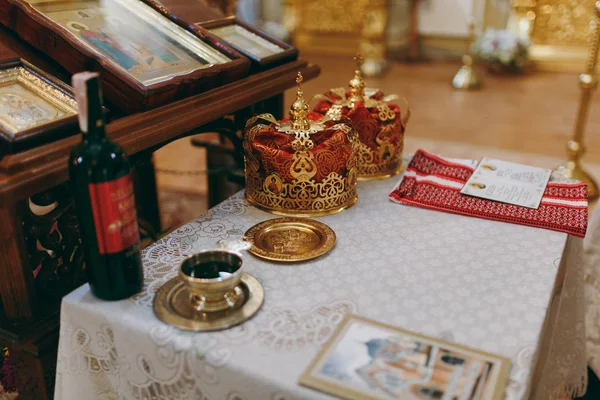Свадьба в православной церкви. Две церемониальные короны, бутылка Кахора, золотая тарелка с обручальными кольцами, святая вода в золотой чаше и другие необходимые предметы на столе рядом с иконостасом — стоковое фото