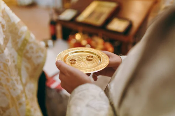 Esküvő az ortodox egyházban. Arany mintás csészealj, jegygyűrű, a menyasszony és a vőlegény a kezében az ember ellen, a háttérben az ikonosztáz és a templombelső — Stock Fotó