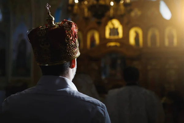 Свадьба в православной церкви. Церемониальная корона на голове жениха в белой рубашке, стоящей позади священника в рясе на темном фоне церковного интерьера — стоковое фото