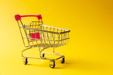 Süpermarket bakkal itme sepetinin siyah jantlar ve tutamacı üzerinde sarı arka plan izole kırmızı plastik öğelerle birlikte alışveriş için kapatın. Alışveriş kavramı. Kopya alanı reklam için