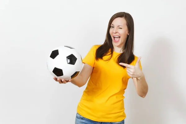 Hermosa joven europea alegre mujer feliz, fanático del fútbol o jugador en uniforme amarillo apuntando a la pelota de fútbol aislado sobre fondo blanco. Deporte, fútbol, salud, concepto de estilo de vida saludable . — Foto de Stock