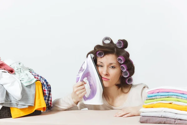 Distressed housewife, curlers on hair in light clothes ironing family clothing on ironing board, put iron to face. Woman isolated on white background. Housekeeping concept. Copy space advertisement. — Stock Photo, Image