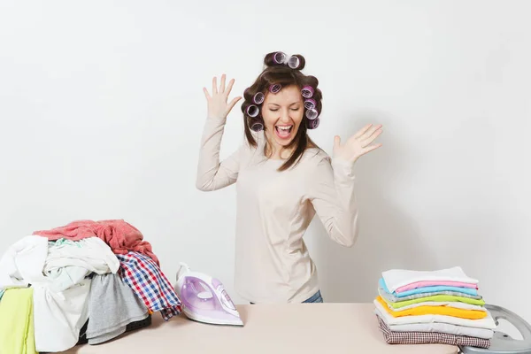 Giovane bella casalinga con bigodini sui capelli in abiti leggeri ballare con abbigliamento di famiglia su asse da stiro con ferro da stiro. Donna isolata su sfondo bianco. Concetto di pulizia Copia spazio annuncio . — Foto Stock