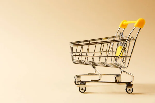 Primer plano de supermercado carrito de compras empuje con ruedas negras y elementos de plástico amarillo en mango aislado sobre fondo beige. Concepto de compras. Copiar espacio para publicidad — Foto de Stock