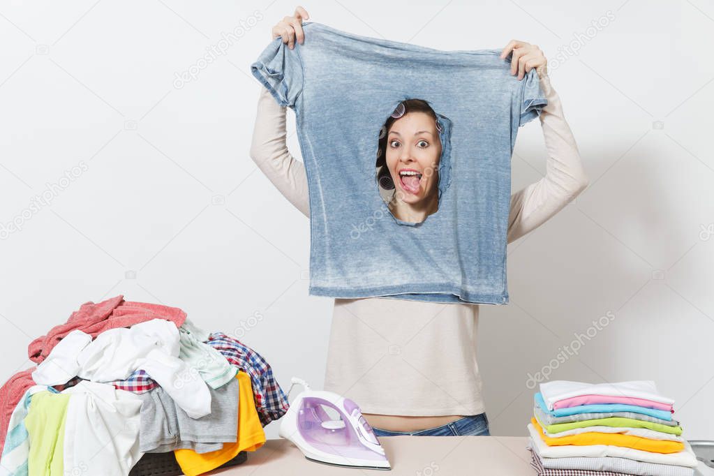Smiling housewife, curlers on hair in light clothes holding burned shirt, looking through hole made by iron standing at ironing board. Woman isolated on white background. Copy space for advertisement.