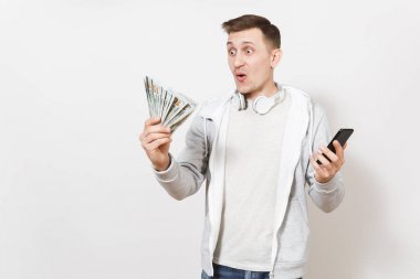 Genç yakışıklı sürpriz öğrenci tişört ve boynuna kulaklıklarla ışık kazak para tutar ve Studio beyaz arka plan üzerinde hareket eden telefon içinde eller. Başarı kavramı
