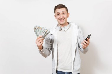 Genç yakışıklı gülümseyen adam öğrenci tişört, beyaz arka plan üzerinde izole elinde bohça-in dolar, nakit para, cep telefonu boynuna kulaklıklarla ışık kazak tutar. Başarı kavramı.