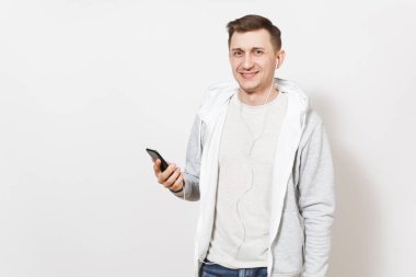 Genç yakışıklı gülümseyen adam öğrenci tişört, kot ve beyaz arka plan üzerinde stüdyoda bir cep telefonunda kulaklık müzik dinlerken ışık kazak. Duygular, iyi ruh kavramı