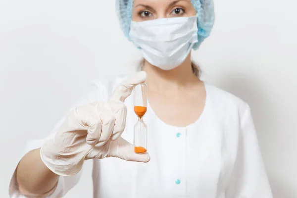 Giovane dottoressa con maschera viso, cappello sterile, guanti isolati su sfondo bianco. Medico chirurgo donna in abito medico tiene clessidra. Personale sanitario, concetto di medicina. Il tempo sta per scadere . — Foto Stock