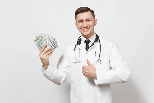 Un jeune médecin souriant isolé sur fond blanc. Médecin masculin en uniforme médical montrant pouces levés, tenant paquet de billets de dollars argent comptant. Personnel de santé, santé, concept de médecine . — Photo