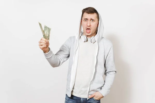 Jonge knappe student in spijkerbroek, t-shirt en lichte sweatshirt met capuchon met koptelefoon houdt twee één-dollarbiljetten en verbaasde blikken in studio op een witte achtergrond. Concept van emoties — Stockfoto