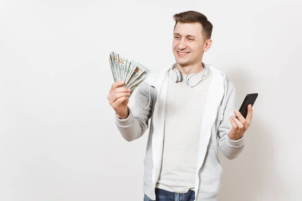 Młody przystojny mężczyzna uśmiechający się w koszulkę i lekka bluza z słuchawek wokół szyi posiada pakiet dolarów, pieniądze i zdejmowany telefon w ręce na białym tle. Koncepcja sukcesu — Zdjęcie stockowe
