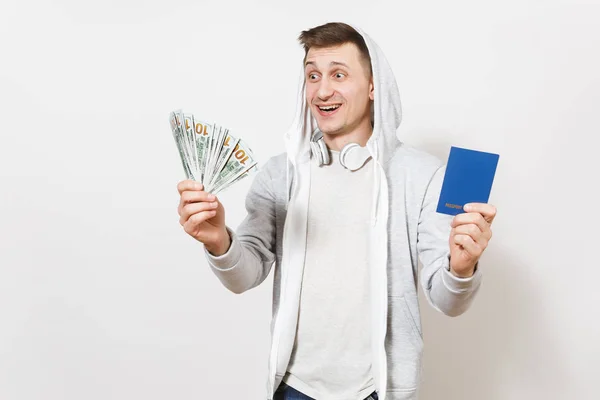 Jeune beau mâle souriant en t-shirt, sweat-shirt léger avec capuche, casque détient passeport international, paquet de dollars, argent liquide isolé sur fond blanc. Concept de voyage, tourisme — Photo