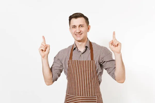 Молодой человек шеф-повар или официант в полосатом коричневом фартуке, рубашка указывает пальцем вверх на копировальное пространство, изолированное на белом фоне. Мужчина-экономка или домработница, смотрящая в камеру. Отечественный работник для рекламы — стоковое фото
