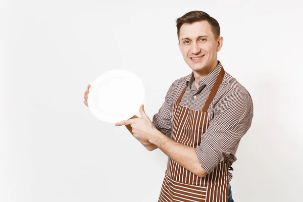 Молодой человек шеф-повар или официант в полосатом коричневом фартуке, рубашка с белой круглой пустой прозрачной тарелкой на белом фоне. Домработница или домработница. Отечественный работник копирует пространство для рекламы — стоковое фото
