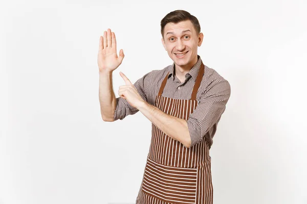 Young smiling man chef or waiter in striped brown apron, shirt showing and pointing on palm to camera isolated on white background. Male housekeeper or houseworker. Domestic worker with copy space. — Stock Photo, Image