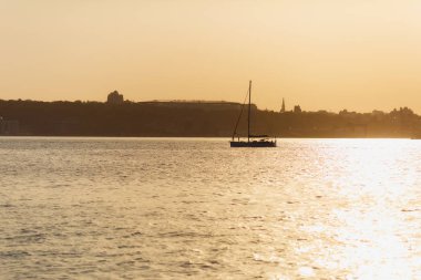 Güzel deniz manzarası. Yalnız arka planı açık sıcak akşam günbatımı sahil şeridi üzerinde endüstriyel bir liman yakınındaki güneş ışığı yansıması ile sakin bir denizde yelkenli tekne