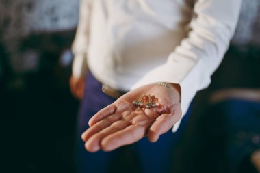 Sabah hazırlık yeni evliler düğün için. Mavi pantolon ve elinde bir bilezik ile beyaz bir gömlek şık bir damat tutar ve parlak bir şekilde dekore edilmiş kol düğmeleri kollu için karanlık bir odada gösterir