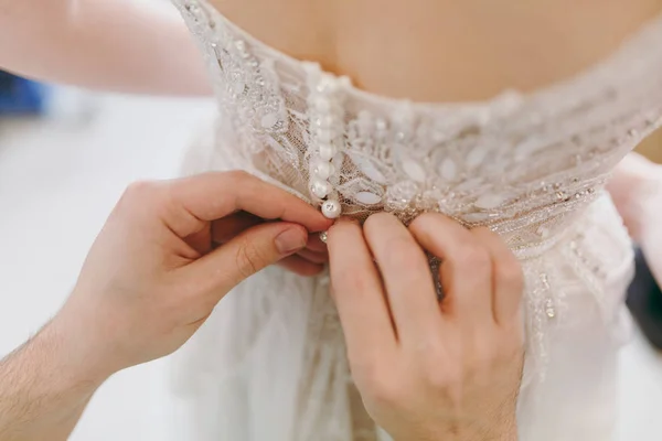 Close up Recém-casados preparação da manhã para o casamento. Mãos masculinas botão de renda elegante delicado vestido de noiva de marfim com contas na cintura para a noiva. Casamento desgaste, acessórios — Fotografia de Stock