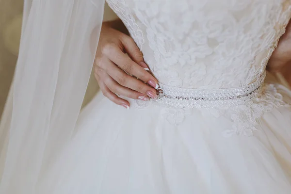 Morning preparation newlyweds for the wedding. Bride in elegant lace white dress with belt with rhinestones holds hand with beautiful patterned manicure on thin waist. Wedding wear, accessories — Stock Photo, Image