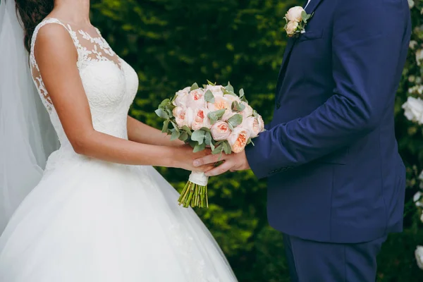 Όμορφη γαμήλια γιορτή. Ο γαμπρός με μπλε κοστούμι με μπουτονιέρα και η νύφη σε μια κομψή δαντέλα φόρεμα με πέπλο και μια ανθοδέσμη παιώνιες και πράσινα φύλλα, κρατώντας τα χέρια στον καταπράσινο κήπο — Φωτογραφία Αρχείου