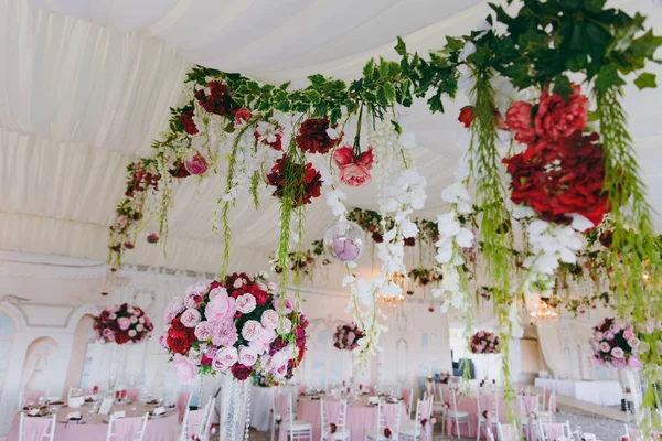Beautiful decoration of the wedding banquet under the awning in pink, burgundy and white tones. Textile ceiling in a banquet hall with flower garlands, ikebans and chandeliers. Wedding organization