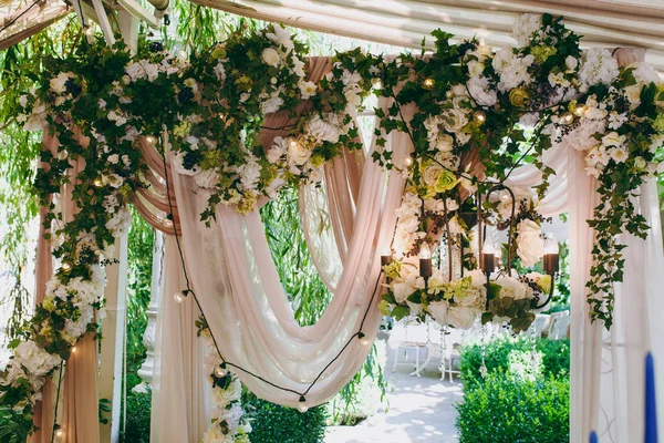 Schöne und exquisite Dekoration der Hochzeitsfeier inmitten eines grünen Gartens. hölzerner rechteckiger Rahmen mit rosa Tuch, vielen hellen Blumen und blauen Beeren dekoriert — Stockfoto