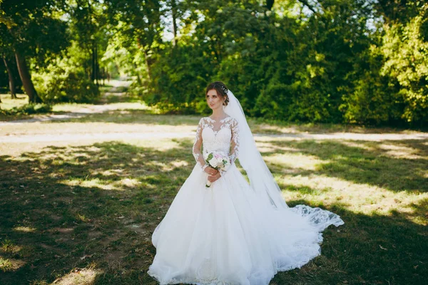 Hermosa sesión de fotos de boda. La novia en un vestido de encaje blanco con una pluma larga, horquilla en el pelo, velo y ramo de flores blancas y rosadas en un gran jardín verde en el día soleado y lluvioso — Foto de Stock