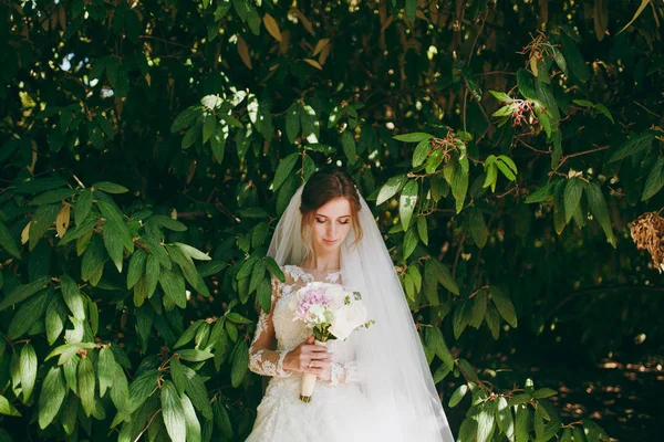 Beautiful wedding photosession. Young tender cute smiling bride in elegant lace dress with long sleeves, veil and bouquet of white and pink flowers amidst the branches of green bush in garden — Stock Photo, Image