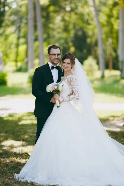 Beautiful wedding photosession. The groom in a black suit and his bride in a white lace dress with a long plume, veil and bouquet smile and embrace in a large green garden on weathery sunny day — Stock Photo, Image
