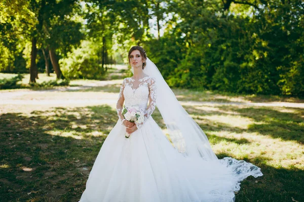 Beautiful wedding photosession. The bride in a white lace dress with a long plume, hairpin in hair, veil and bouquet of white and pink flowers in a large green garden on weathery sunny day — Stock Photo, Image