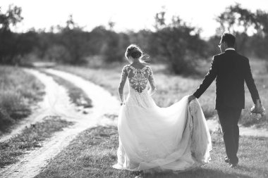 Güzel düğün photosession. Yakışıklı damat bir siyah takım elbise ve dantel elbise büyük alan arka plan ağaç ve çalı etrafında yürüyüş üzerinde zarif saç modeli ile genç gelin. Siyah ve beyaz