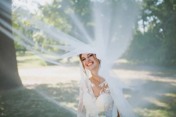 Beautiful wedding photosession. Portrait of the young smiling bride in a white lace dress and bouquet of flowers in a fluttering white veil in a large green garden on weathery sunny day — Stock Photo, Image