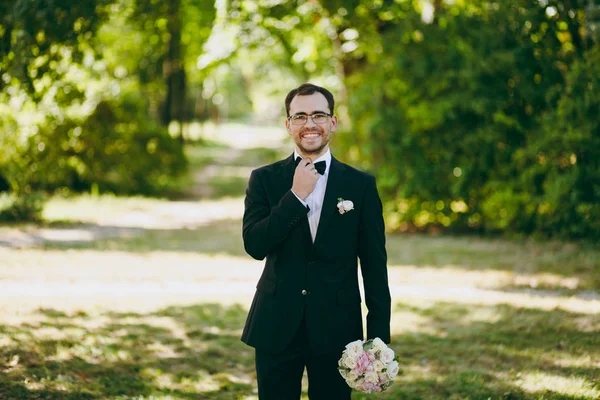 Vackert bröllop fotosession. Brudgummen i svart kostym, vit skjorta och glasögon med bukett i vitt och rosa blommor för bruden rätar ut en fluga i en stor grönskande trädgård weathery solig dag — Stockfoto