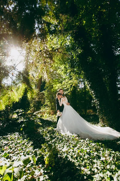 Beautiful wedding photosession. The handsome groom in a black suit, white shirt and glasses with bouquet of flowers and his young cute bride in white lace dress in large green garden on sunny day — Stock Photo, Image