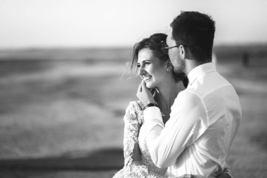 Güzel düğün photosession. Beyaz gömlekli yakışıklı tıraşsız damat yürüyüş kıyı şeridi boyunca mükemmel saç modeli ile dantel desen elbiseli genç sevimli gülümseyen gelini öper. Siyah ve beyaz