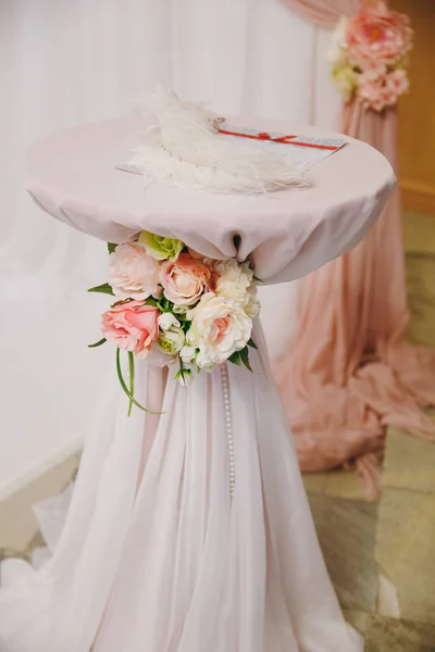 Mesa redonda para pintar decorada con un paño rosa, flores y cuentas en el fondo de un hermoso arco de bodas. Una pluma blanca grande y una carta sobre la mesa. Decoración de la boda, accesorios —  Fotos de Stock