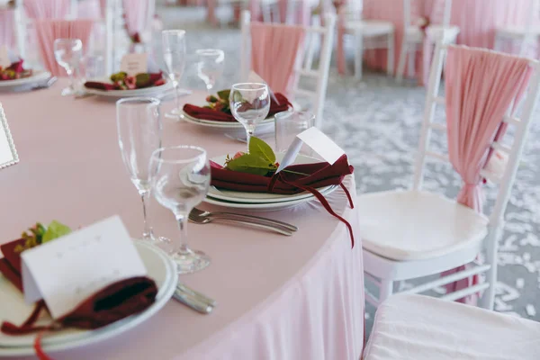 Красива прикраса весільного бенкету під тентом в рожевих, бордових і білих тонах. Обстановка столу в залі з тарілками, столовими приборами, келихами, серветками та картами, прикрашеними квітковими композиціями — стокове фото