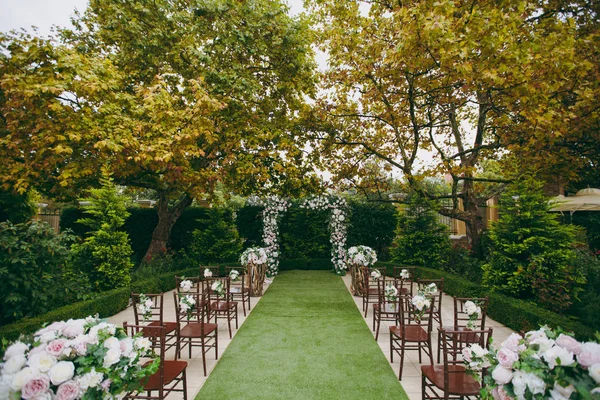 Bela decoração de uma cerimônia de casamento em um jardim de outono verde. Cadeiras de madeira marrom para convidados de ambos os lados da passarela, o que leva a um arco festivo decorado com flores — Fotografia de Stock