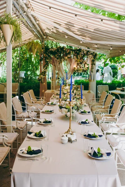 Beautiful and exquisite decoration of the wedding celebration in in the middle of a green garden. Banquet served table with a beige tablecloth, plates and candlesticks with blue candles under cover — Stock Photo, Image