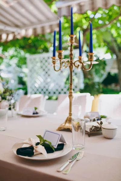 Beautiful and exquisite decoration of the wedding celebration in in in in the middle of a green garden. Банкетный стол с бежевой скатертью, тарелками и подсвечниками с голубыми свечами под крышкой — стоковое фото