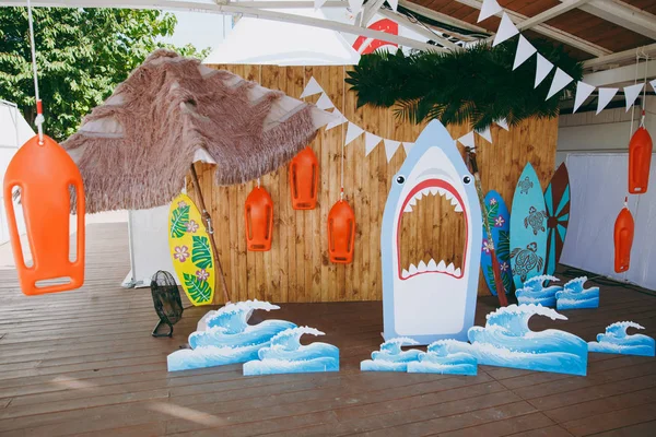 Beautiful scenery and design of a child's birthday party in a marine style with surfers, sharks, waves, garlands in the open air under the awning. Organization and decoration of family holidays